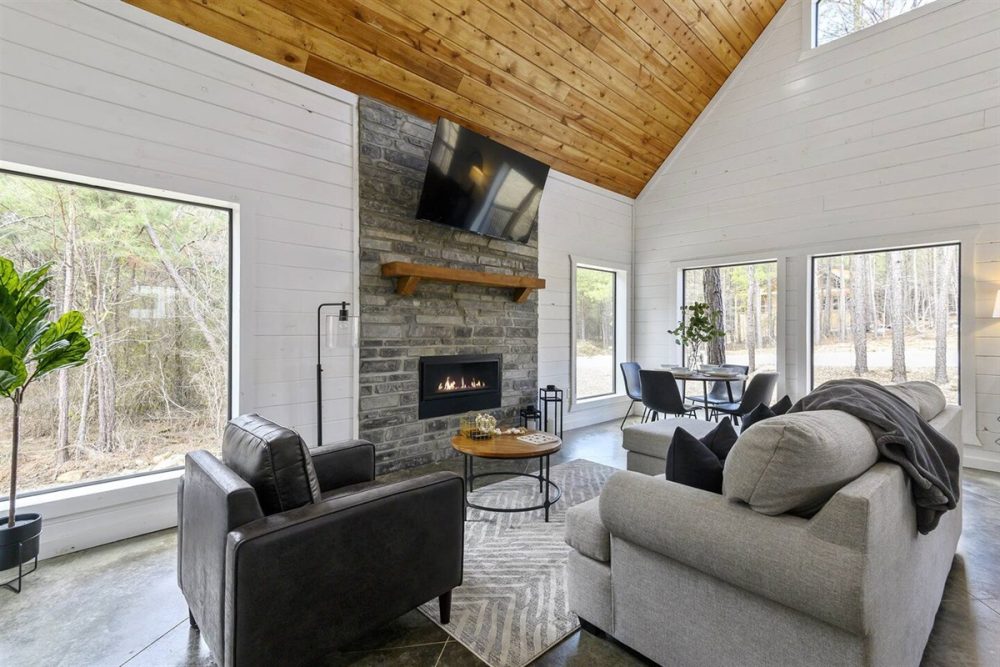 Cozy cabin interior featuring a stone fireplace with a television, comfortable seating, and large windows