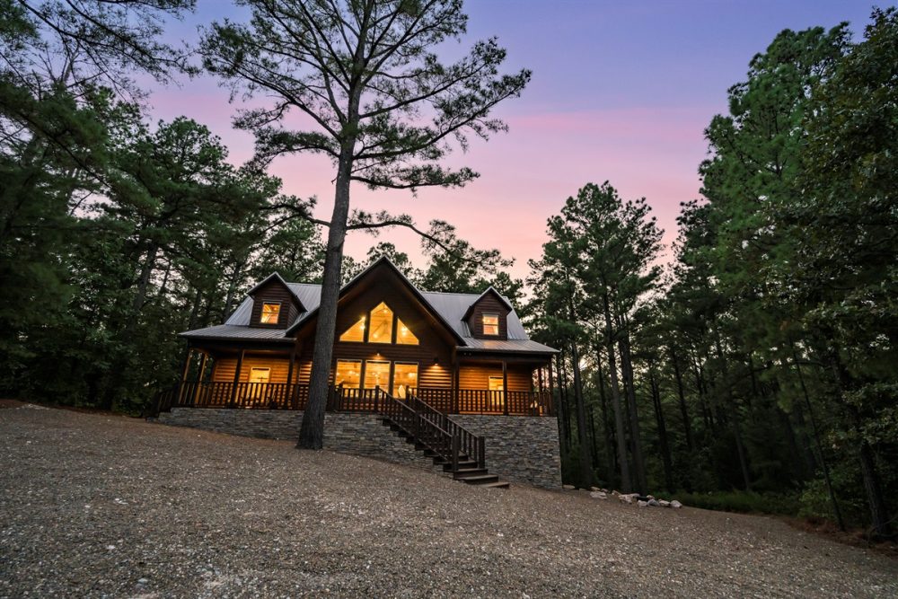 Cozy log cabin surrounded by tall trees at dusk, featuring warm glowing windows